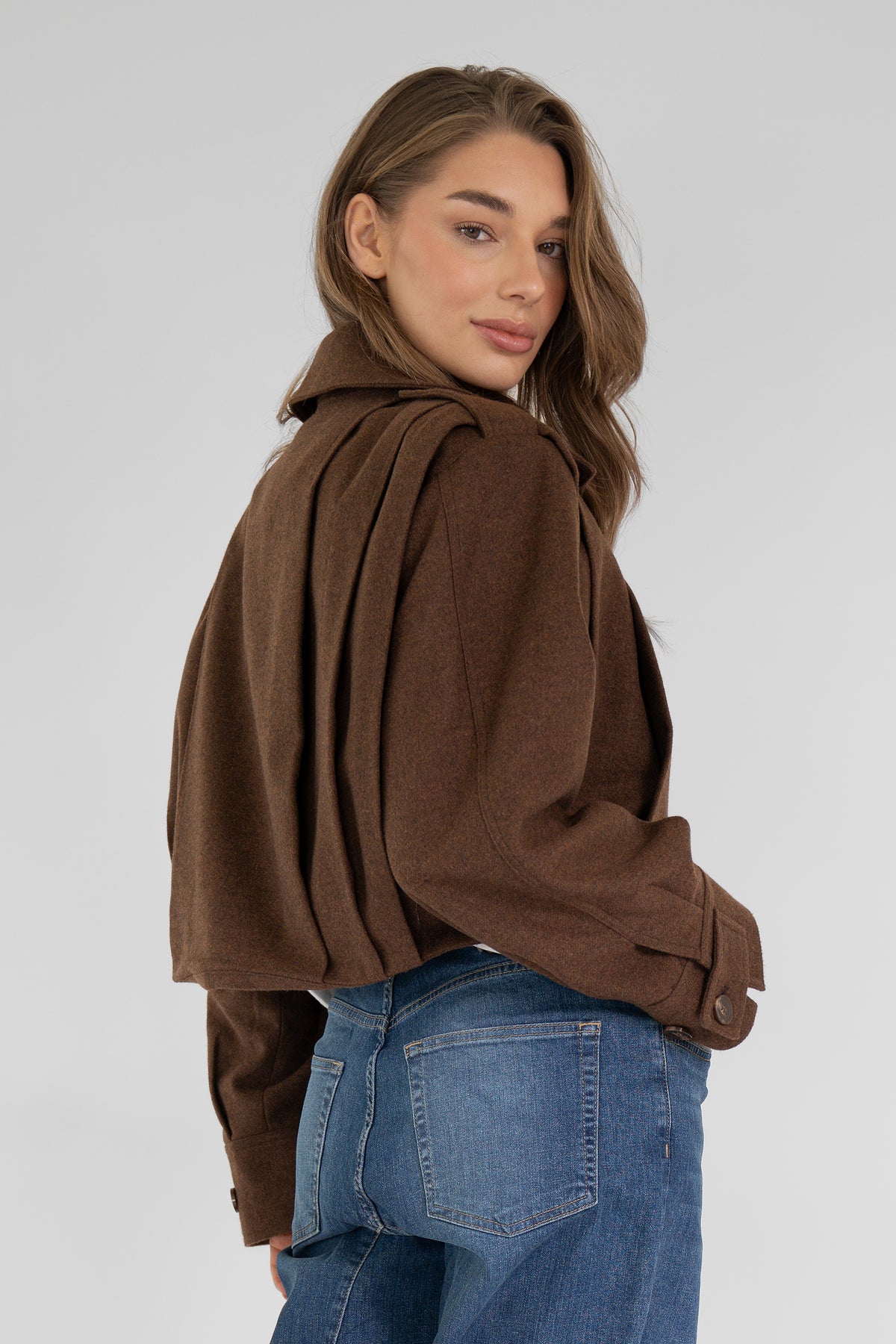Zijkant van de bruine JOSSY JACKET met plooien op de schouder, model kijkt over haar schouder